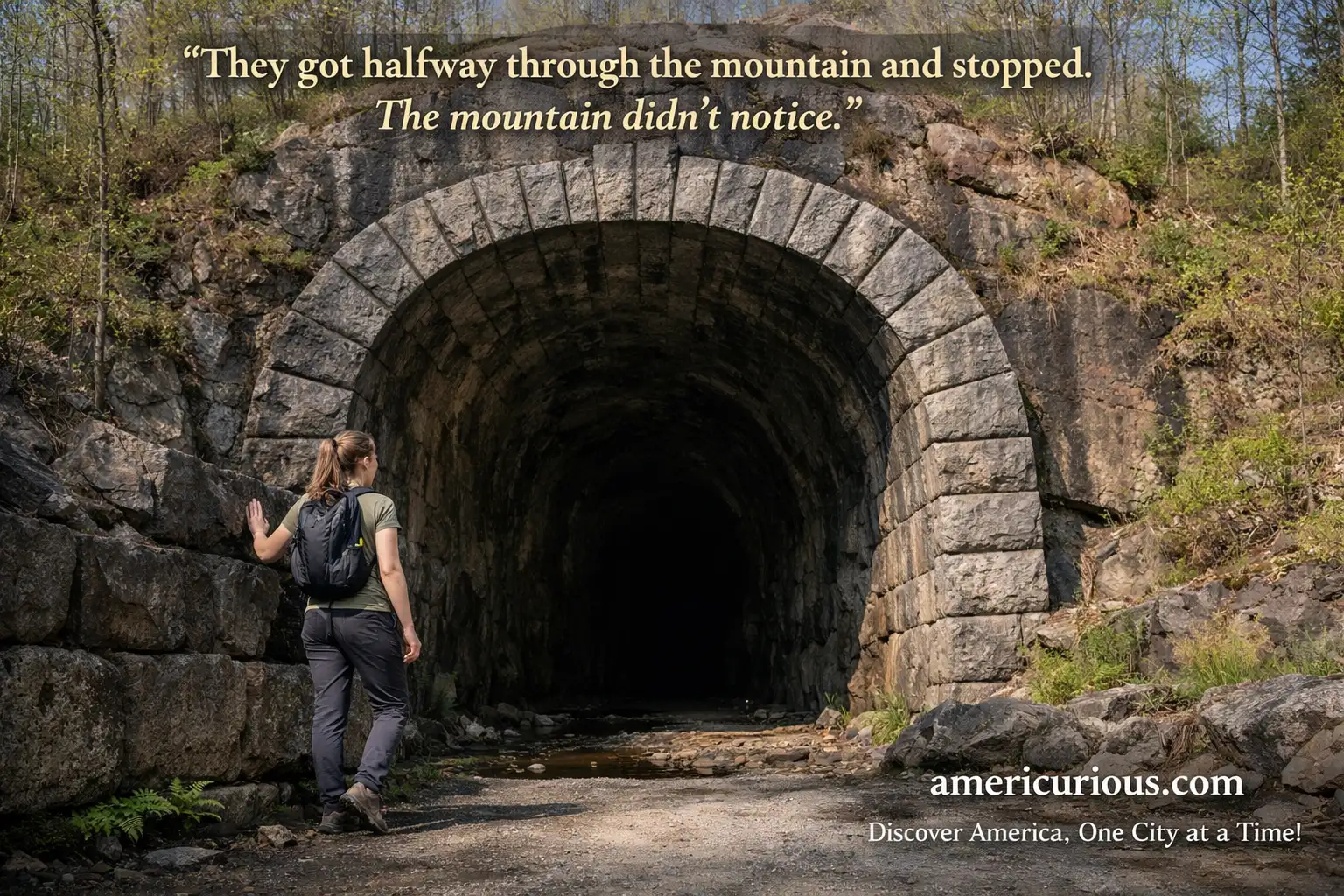 Stone arch entrance of Stumphouse Tunnel in Oconee County, South Carolina, with a woman pausing at the threshold as the hand-drilled 1850s passage recedes into darkness against a forested Blue Ridge hillside. Day trips from Greenville SC, Stumphouse Tunnel Walhalla.